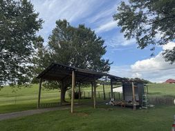 A wooden carport with a tin roof, set on green grass with a tree in the background under a blue sky.