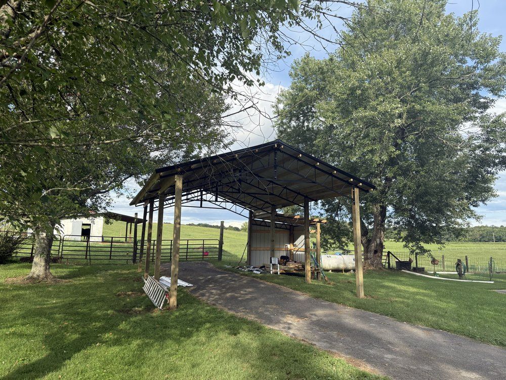 Carport with metal roof supported by wooden posts, driveway leading to it, and green pasture in background.