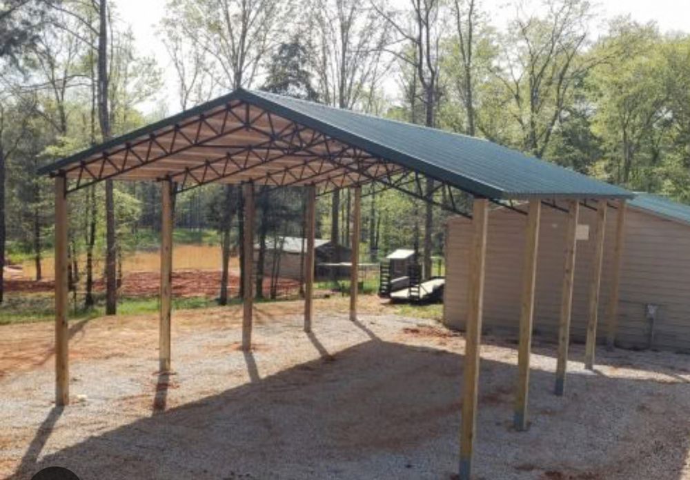 A carport with a green roof, supported by wooden posts, in a gravel-covered area with trees in the background.