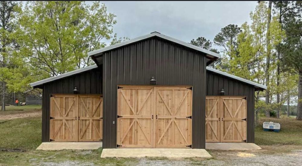 Brown metal barn with three wooden doors. Green trees in the background, overcast sky.