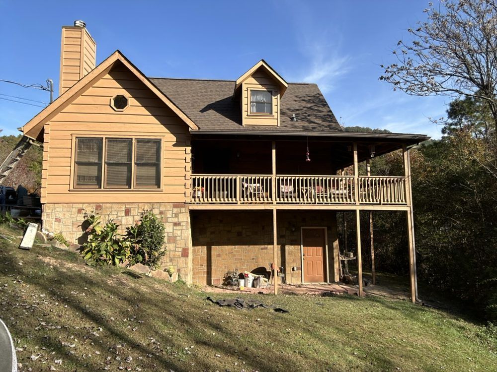 Brown cabin on a hillside with a porch and stone foundation. Blue sky.