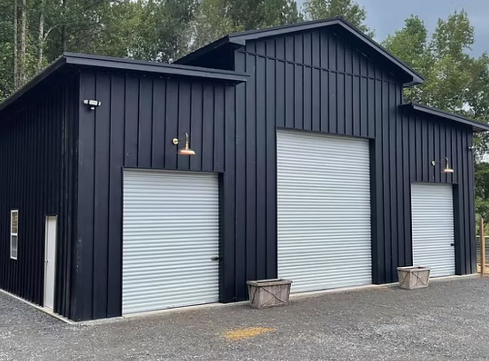 Black metal barn with three white garage doors and exterior lights, set on a gravel driveway.