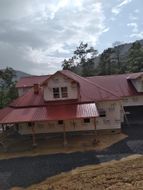 Red-roofed house under construction; white siding, windows, and porch with wooden posts. Mountain backdrop on a cloudy day.
