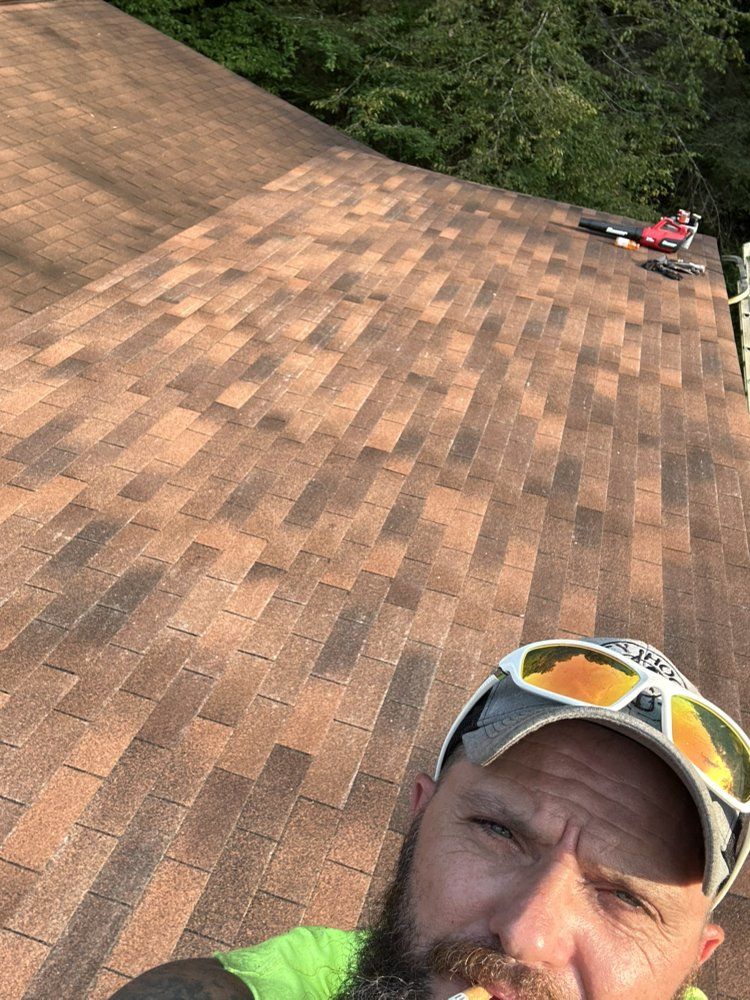 Man on roof with sunglasses, brown shingles, tools, and trees in background.