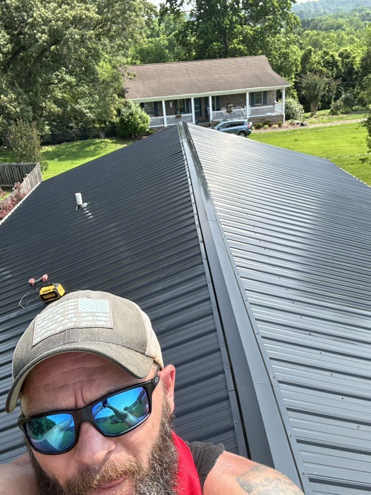 Man on metal roof, selfie. Wearing hat, sunglasses, red strap. House and trees in background.