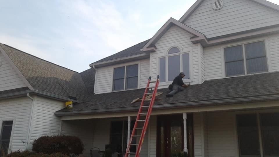 Person on a ladder cleaning roof gutters of a white house with dark gray shingles.