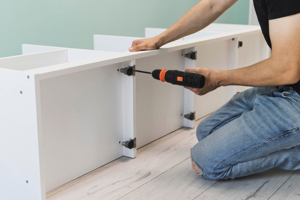 A person uses a cordless drill to attach a vertical support to a white wooden shelf unit on a light-colored floor.