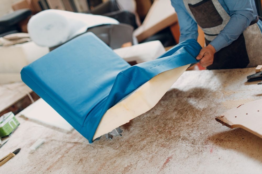 A person wearing an apron upholsters a cushion with bright blue fabric in a workshop.