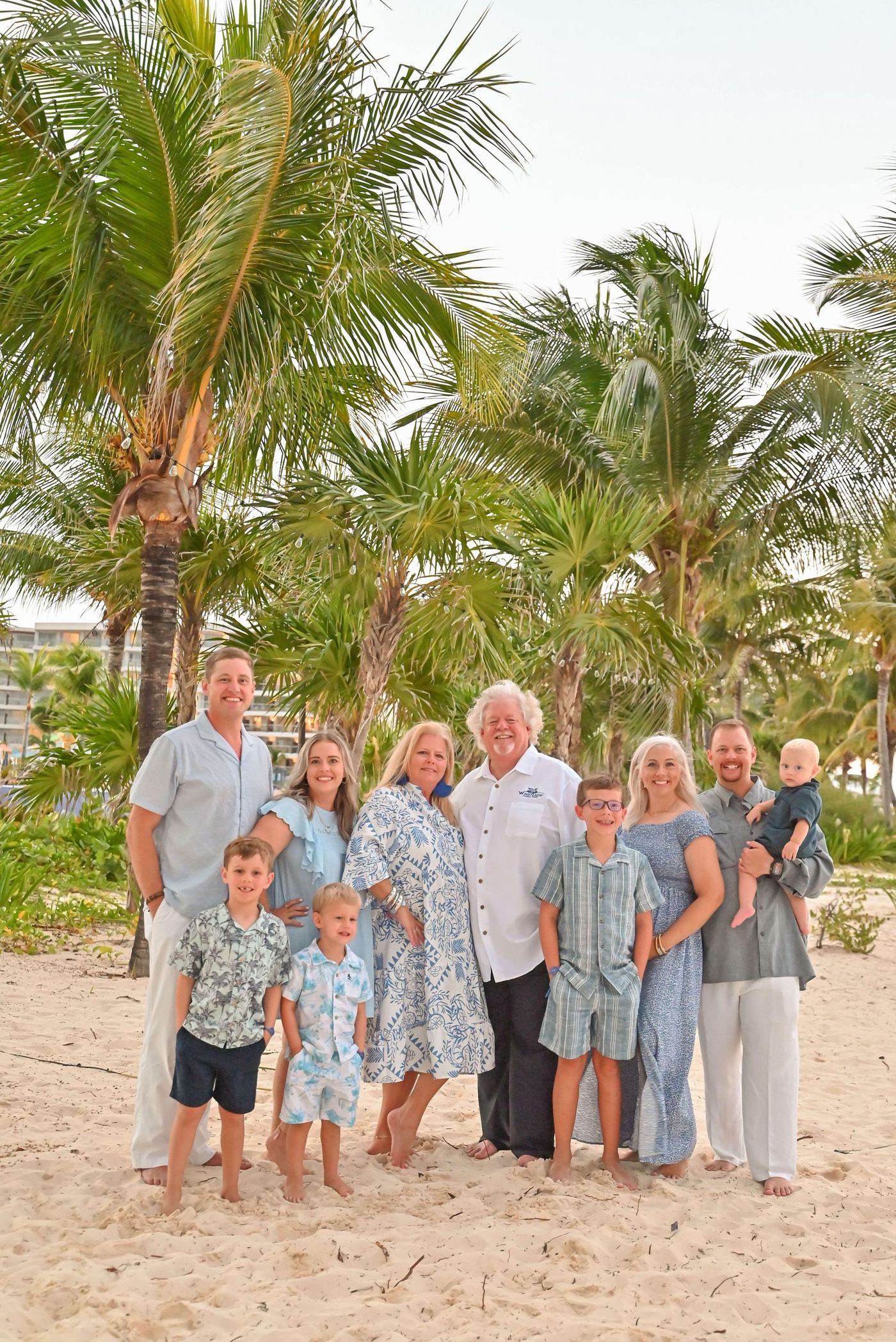 A multi-generational family stands together on a wooden boardwalk at a beach with palm trees in the background.