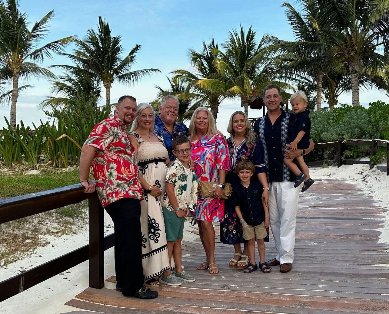 A multi-generational family stands together on a wooden boardwalk at a beach with palm trees in the background.