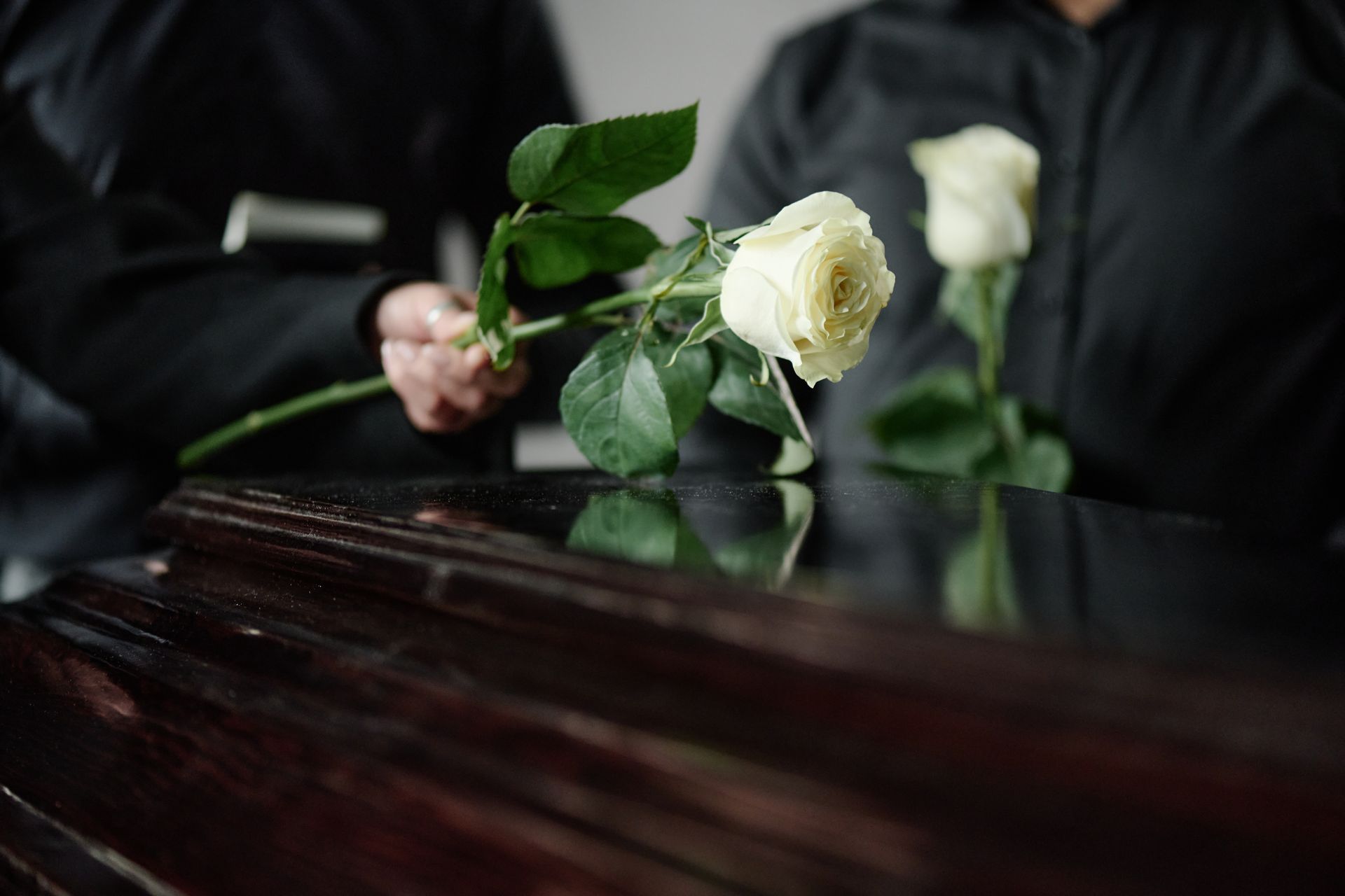 Two people in black clothing holding white roses over a dark wooden coffin.