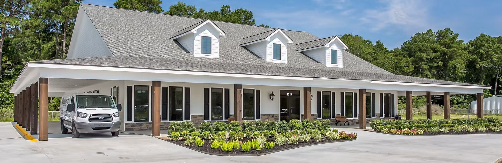 White building with three dormers, carport, and a van in front, surrounded by trees.
