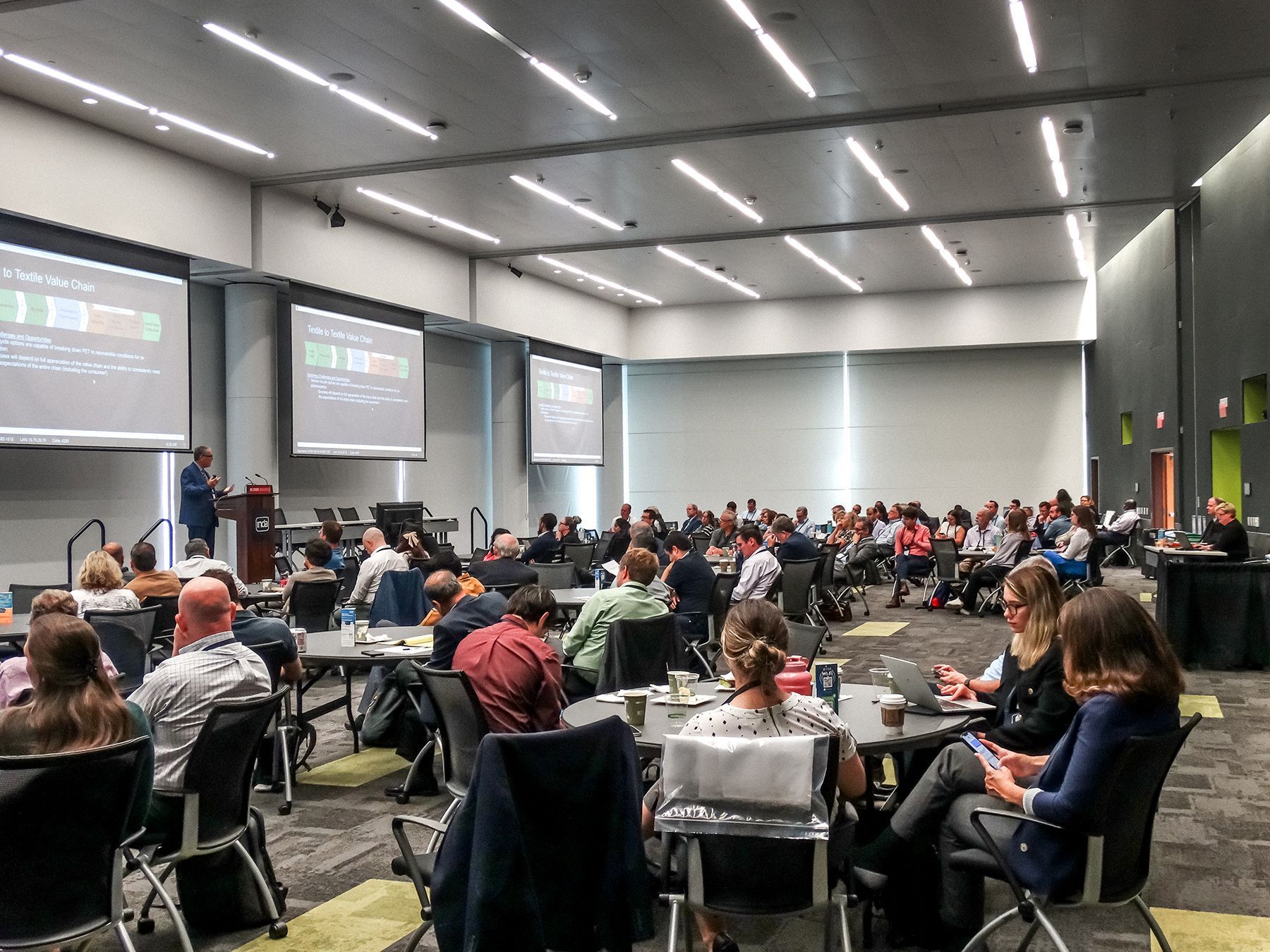 Conference room with speaker presenting to seated audience; screens show graphs.