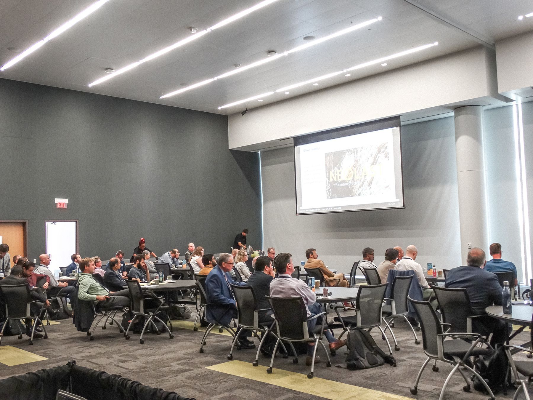 Conference room with people seated at tables, watching a presentation on a screen.