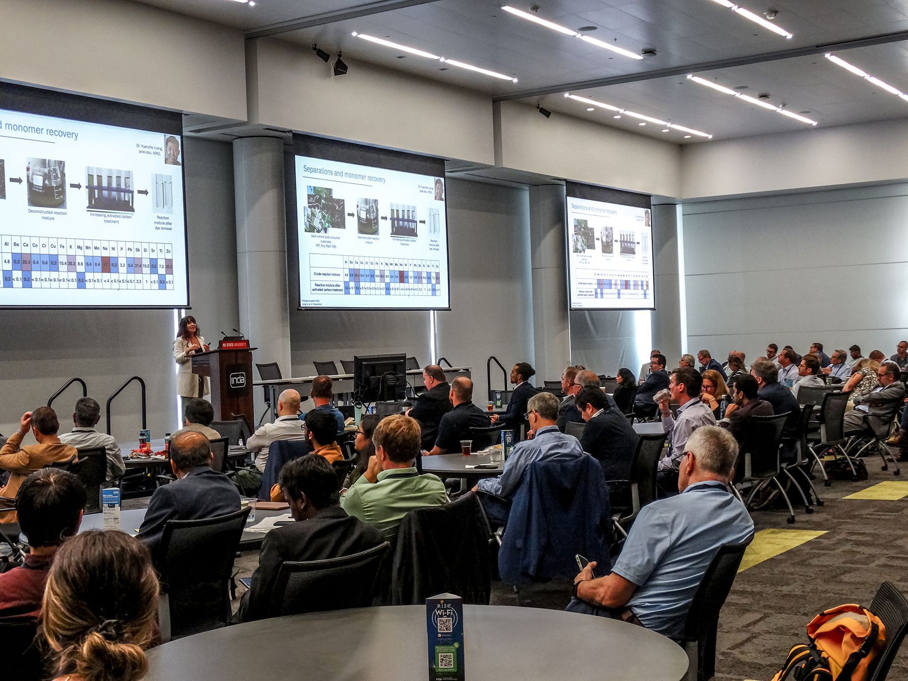 Woman presenting at conference; large screens with diagrams, audience seated at tables.