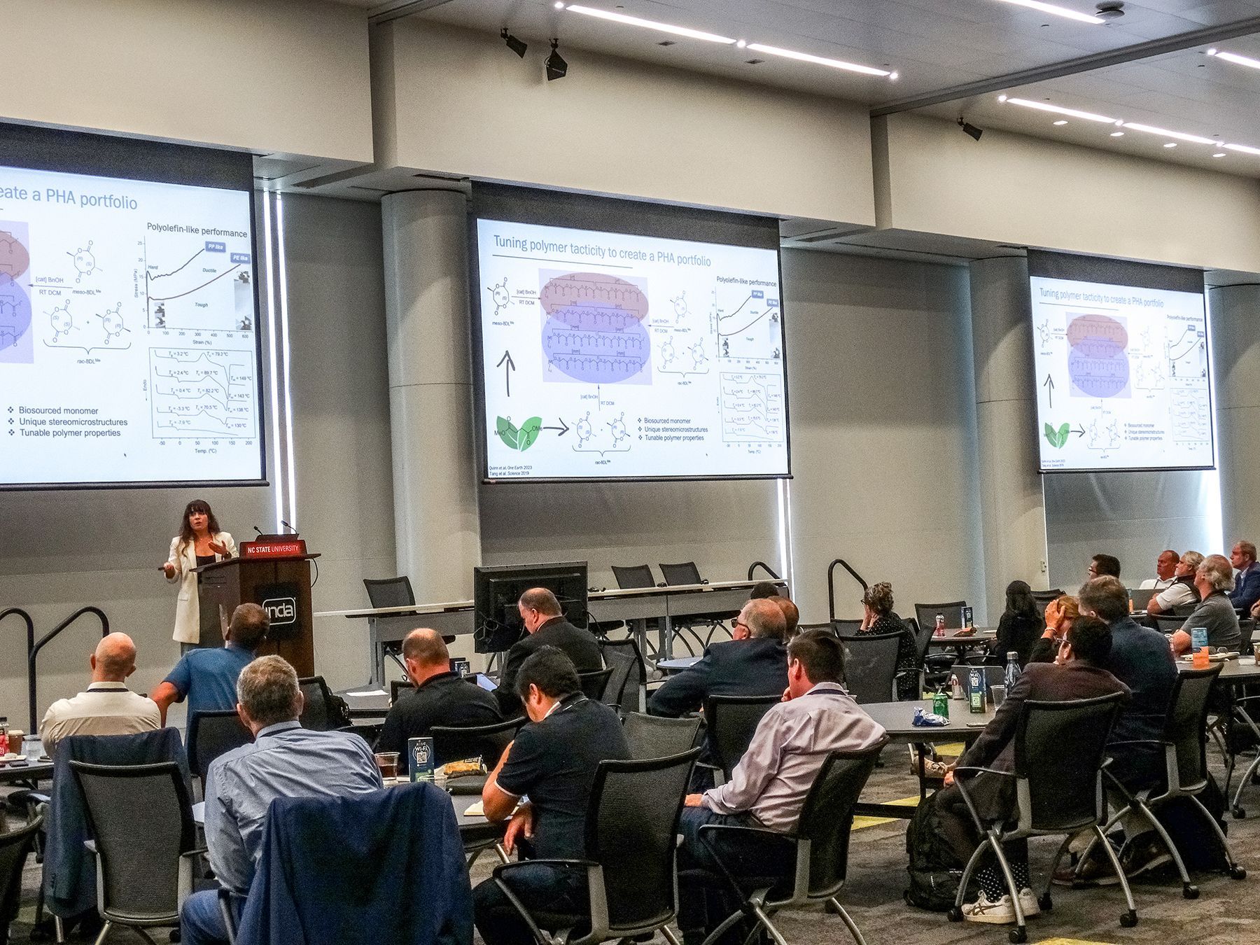 Woman presenting to a seated audience with three screens showing diagrams. Conference hall.
