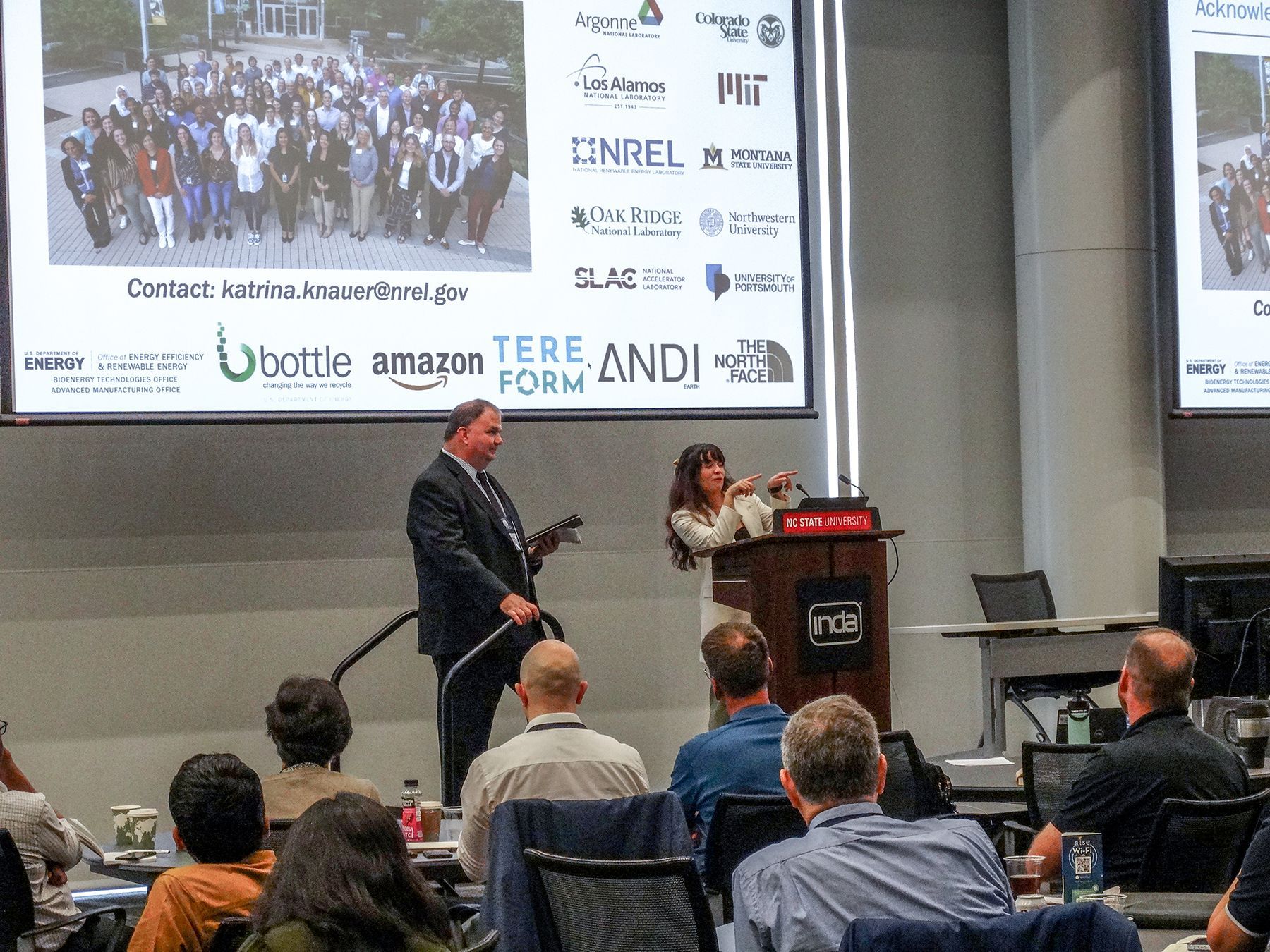 Man and woman on stage presenting; audience watching. Presentation screen, logos visible.