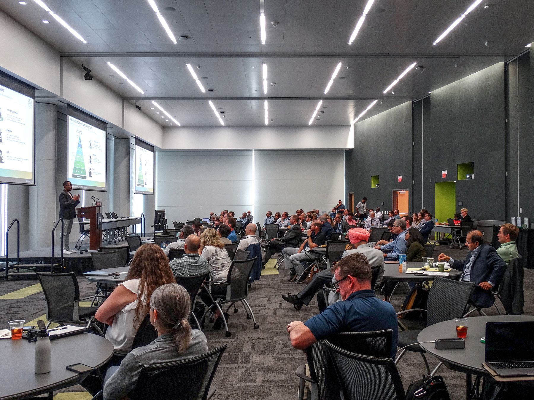 Conference presentation in a large hall with audience seated at tables, listening to a speaker.