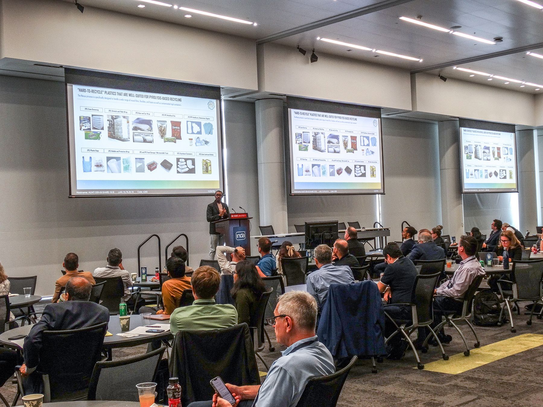 A speaker presents to a seated audience, three presentation screens behind him.