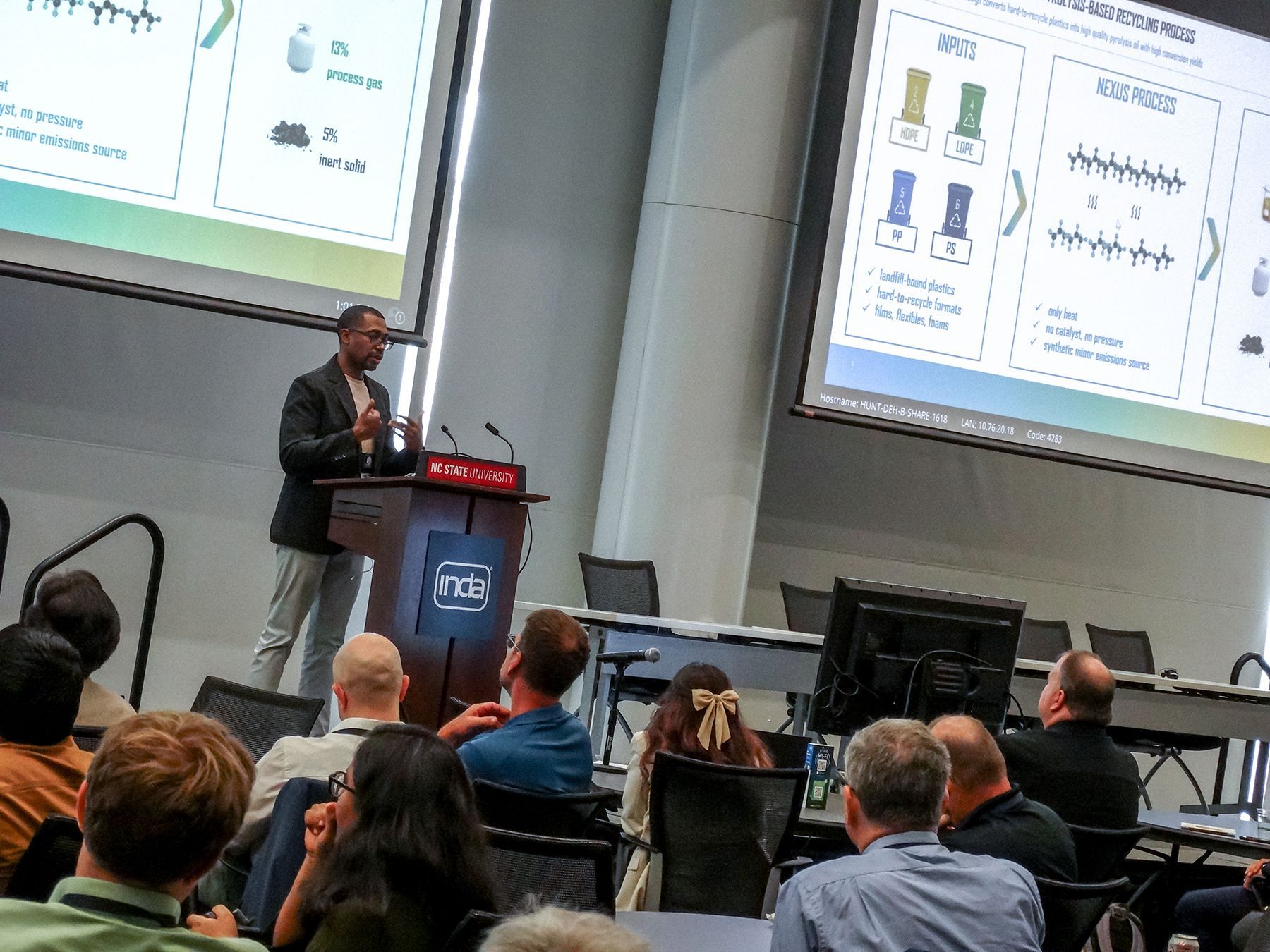 Man presenting at a conference, slides displayed, audience seated, inside a hall.