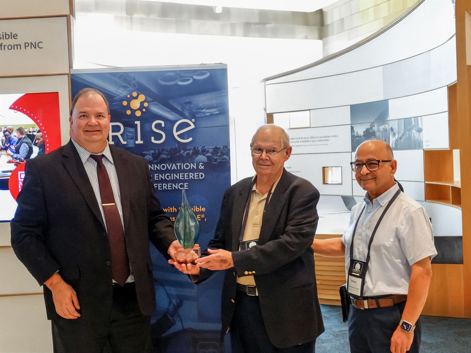 Three men holding an award; one in a suit, two in casual wear; in front of a conference banner.