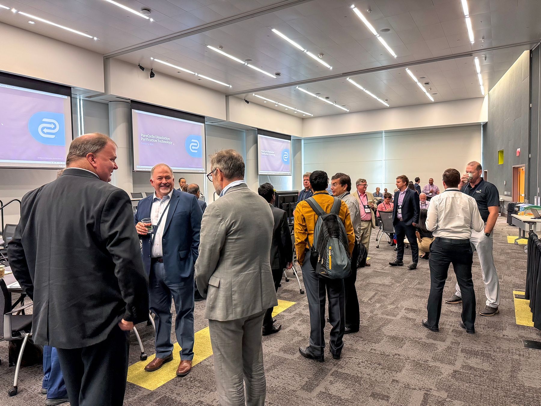 People networking in a modern conference room. Several screens display a logo.