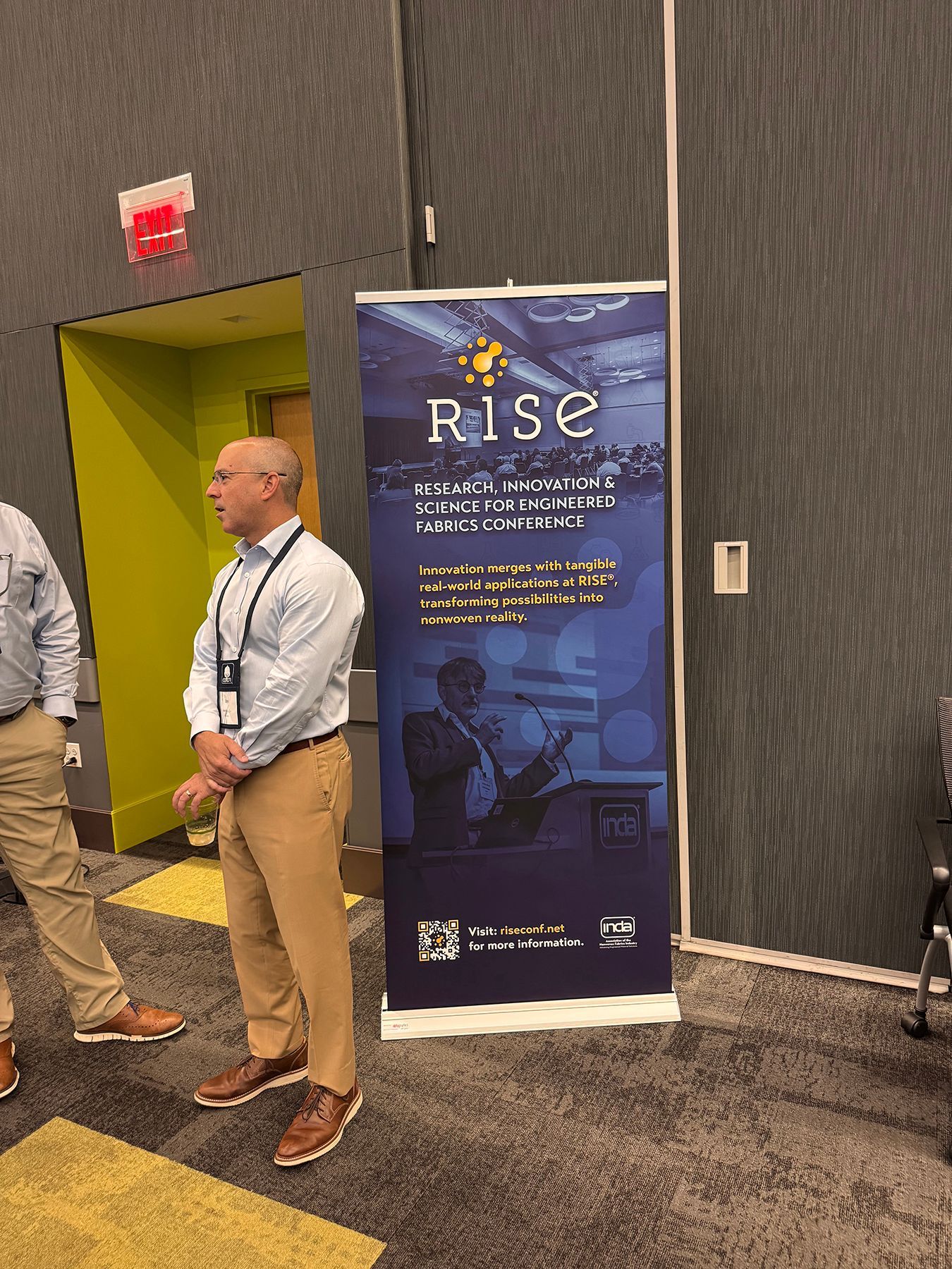 Man standing by a RISE banner in a conference setting. He is wearing a light blue shirt, tan pants, and brown shoes.