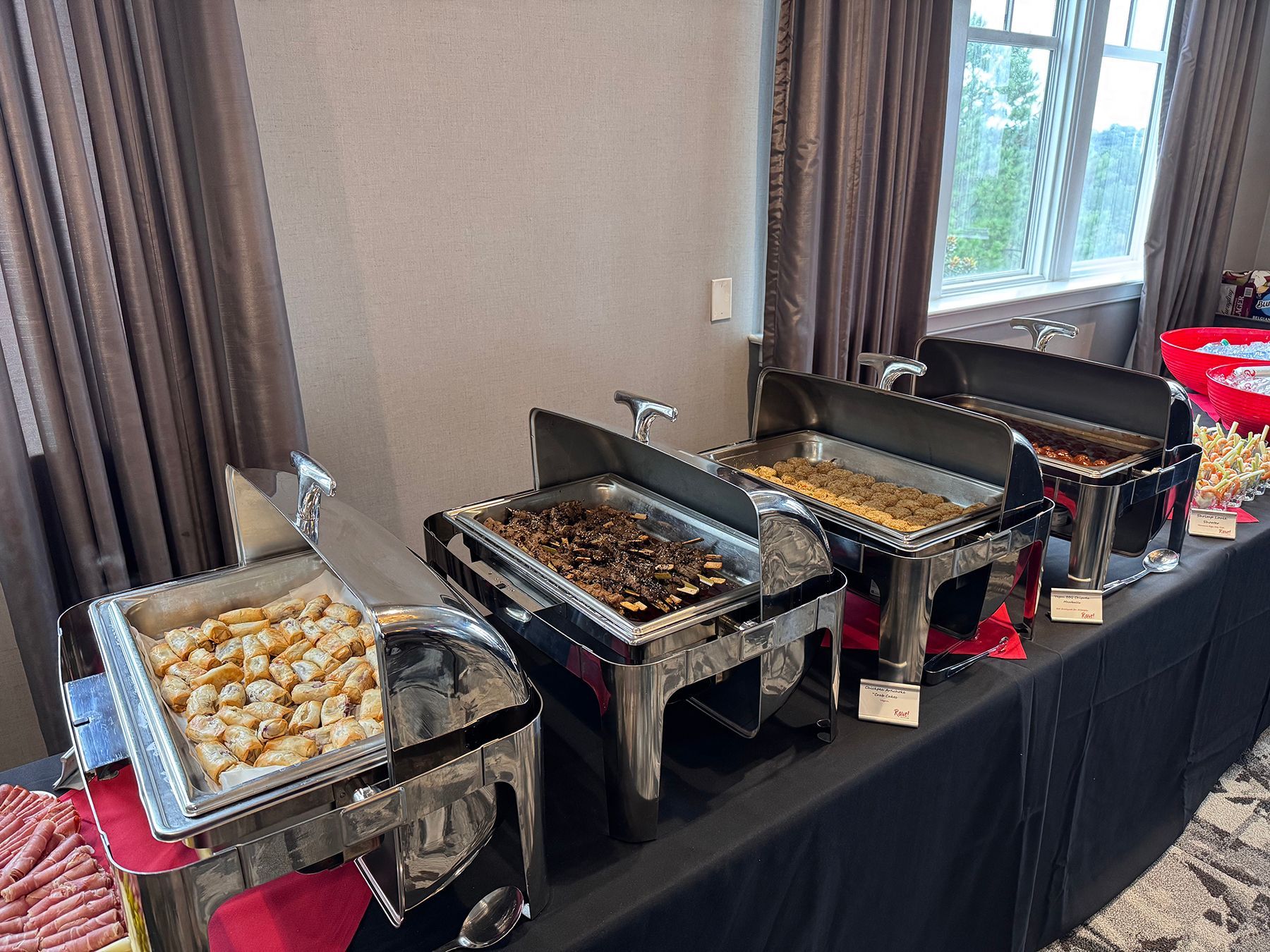 Buffet table with several silver chafing dishes containing food, set in a room with a window and curtains.