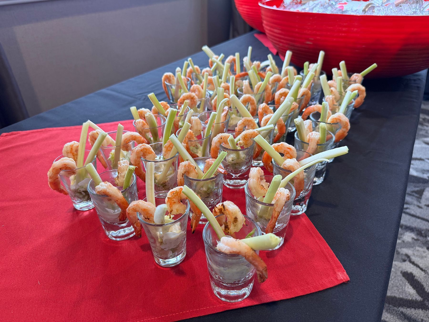 Shrimp cocktail appetizers in shot glasses with celery sticks on a red table.