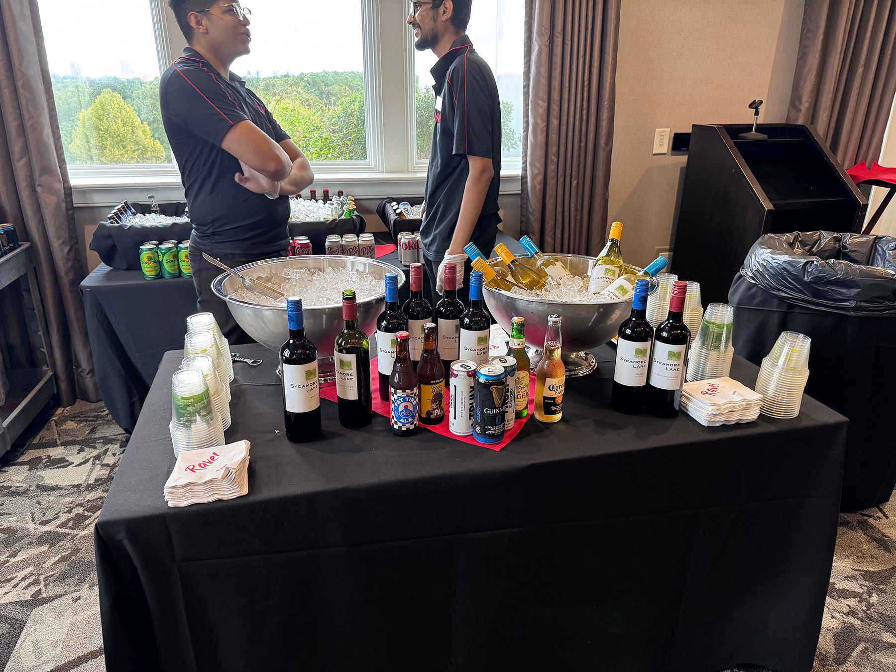 Table with wine bottles and drinks, two men talking by window.