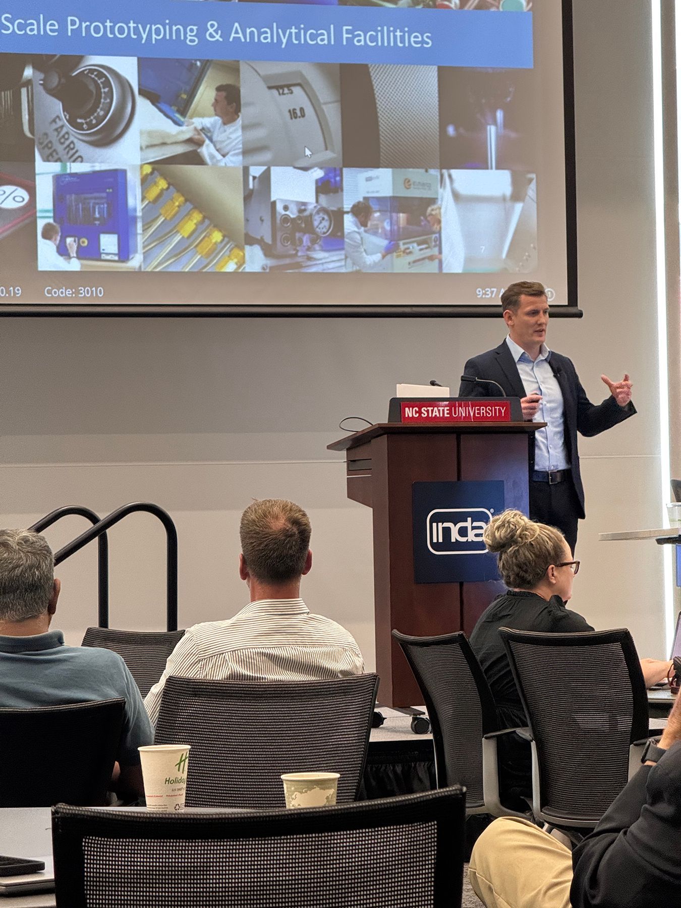 Man presenting at a conference. Audience seated, watching. Large screen in background showing images.
