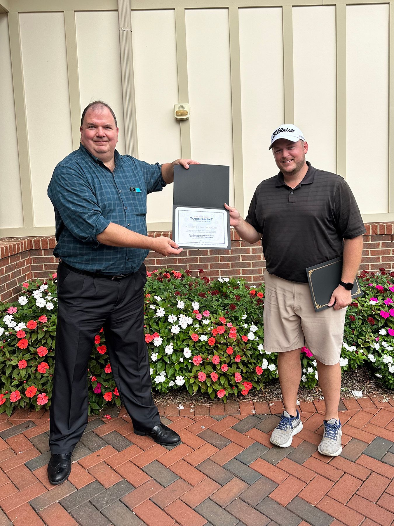 Two men, one holding a certificate, standing by flowers. Outdoors setting, brick and beige wall.