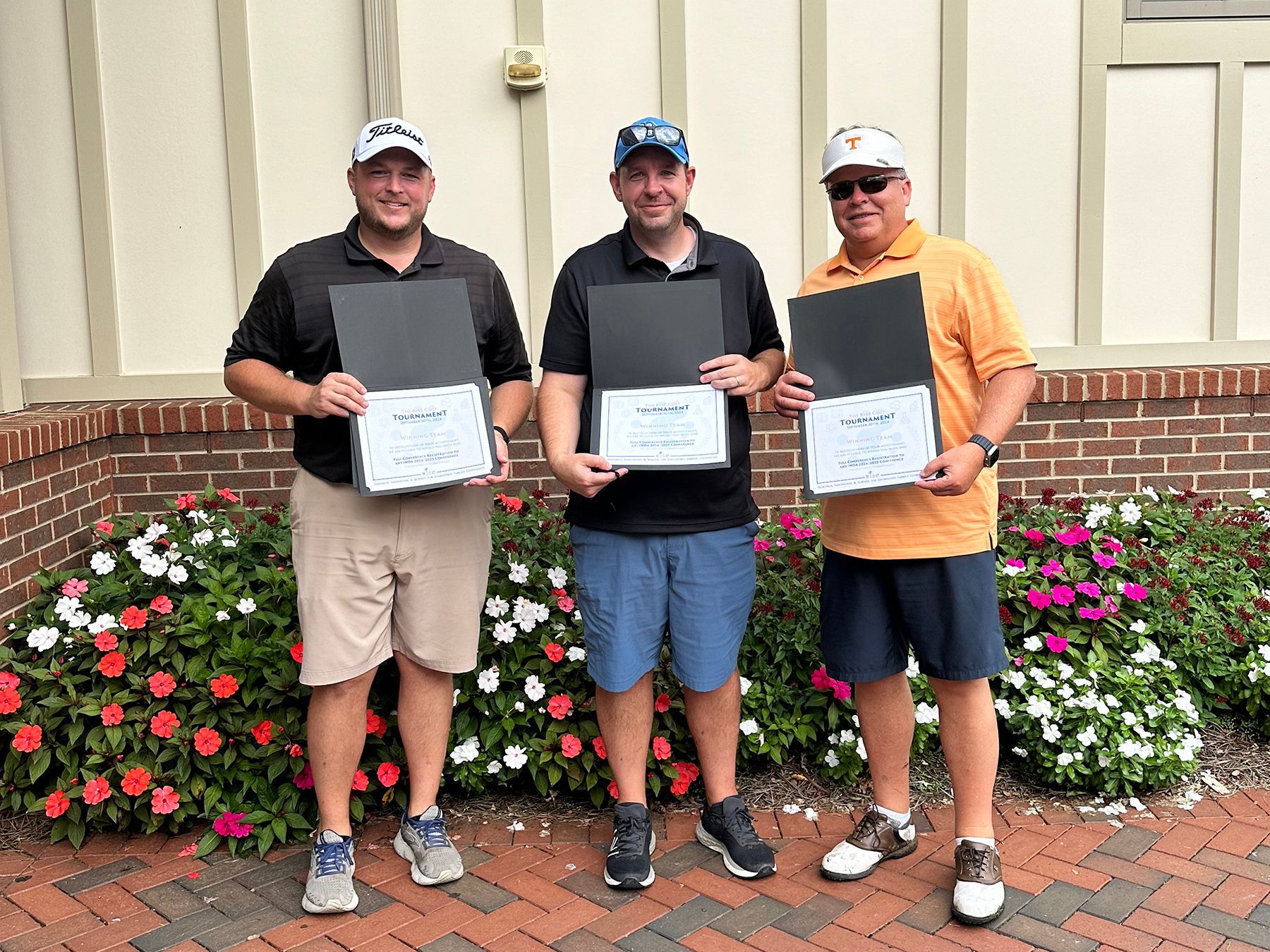Three men holding certificates, standing in front of flowers and a brick building.