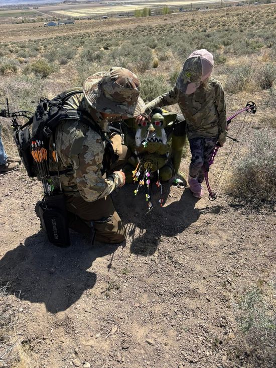 A person in camouflage kneels next to a child, examining something in a field.