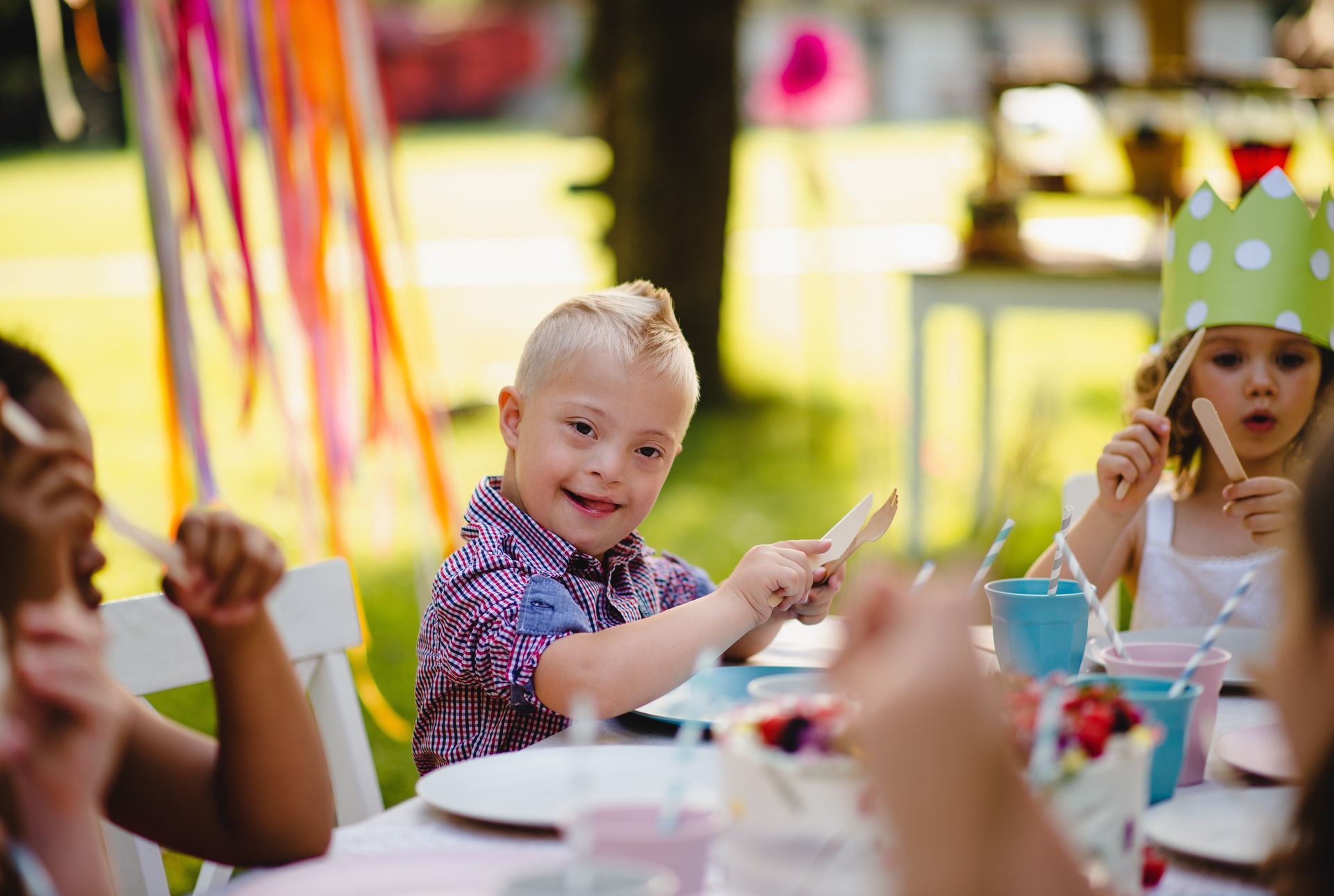 Child with Down syndrome smiles at outdoor party table, holding a wooden spoon.