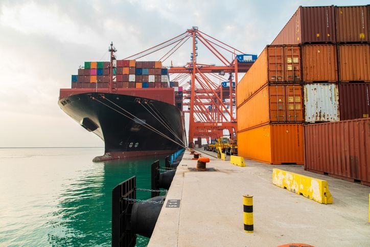 Cargo ship docked at a port with stacked shipping containers and a large crane under a cloudy sky.