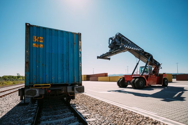 A forklift loading a blue shipping container from a rail car on a sunny day.