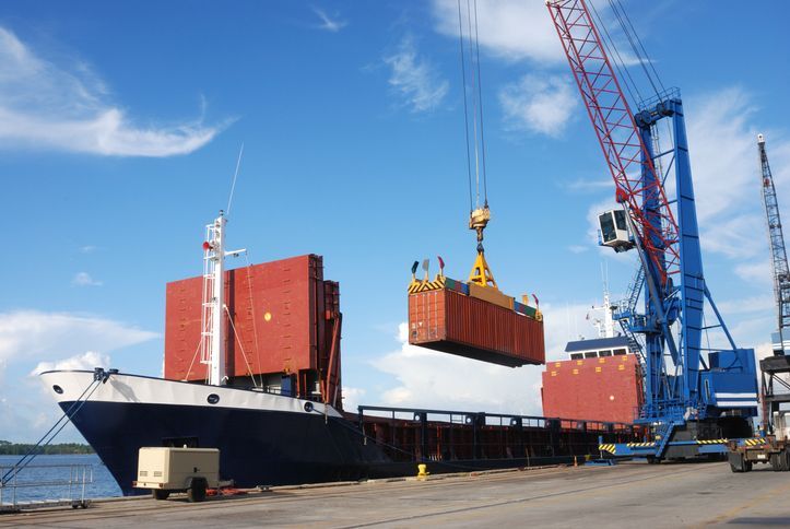 Cargo ship being loaded with a container by a large blue crane at a dock, under a bright blue sky.