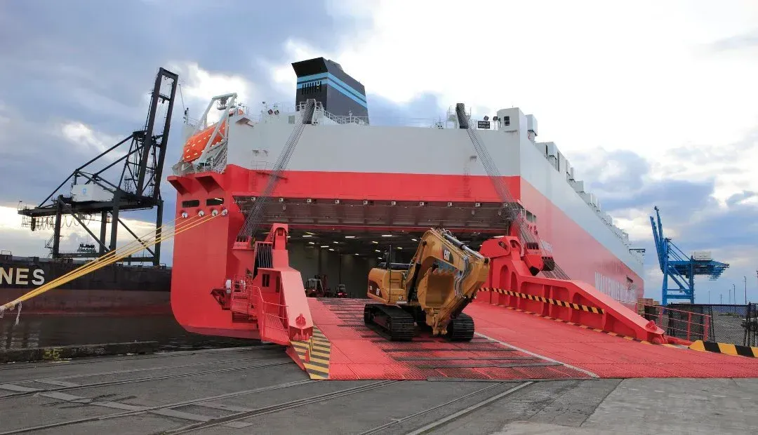 Car carrier truck with several cars on a pier, near water. Buildings and mountains in the background.