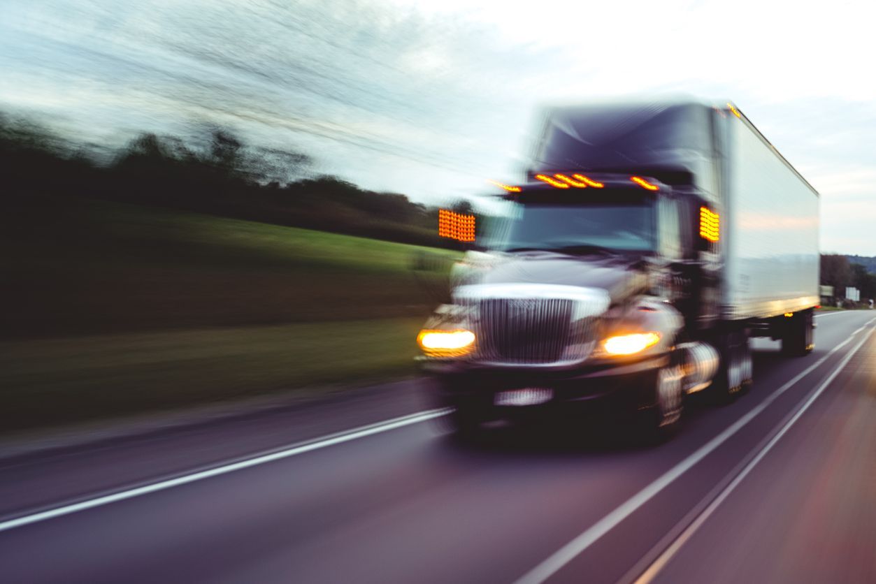 Black semi-truck speeding on highway, motion blur, daytime, cloudy sky, trees in background.
