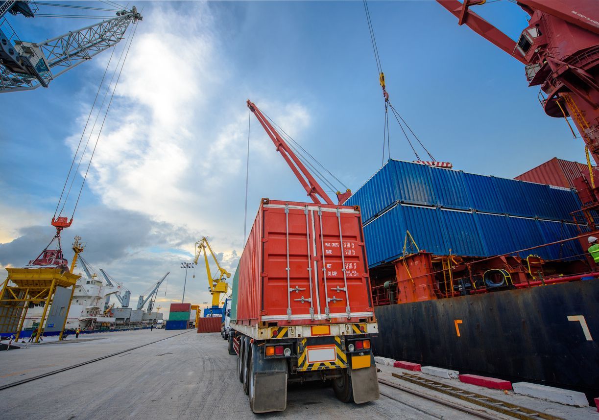 Truck loading cargo container at a busy port with cranes and a large ship.