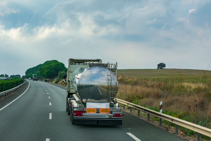Tanker truck driving down a highway with green trees and a field on the side under a cloudy sky.