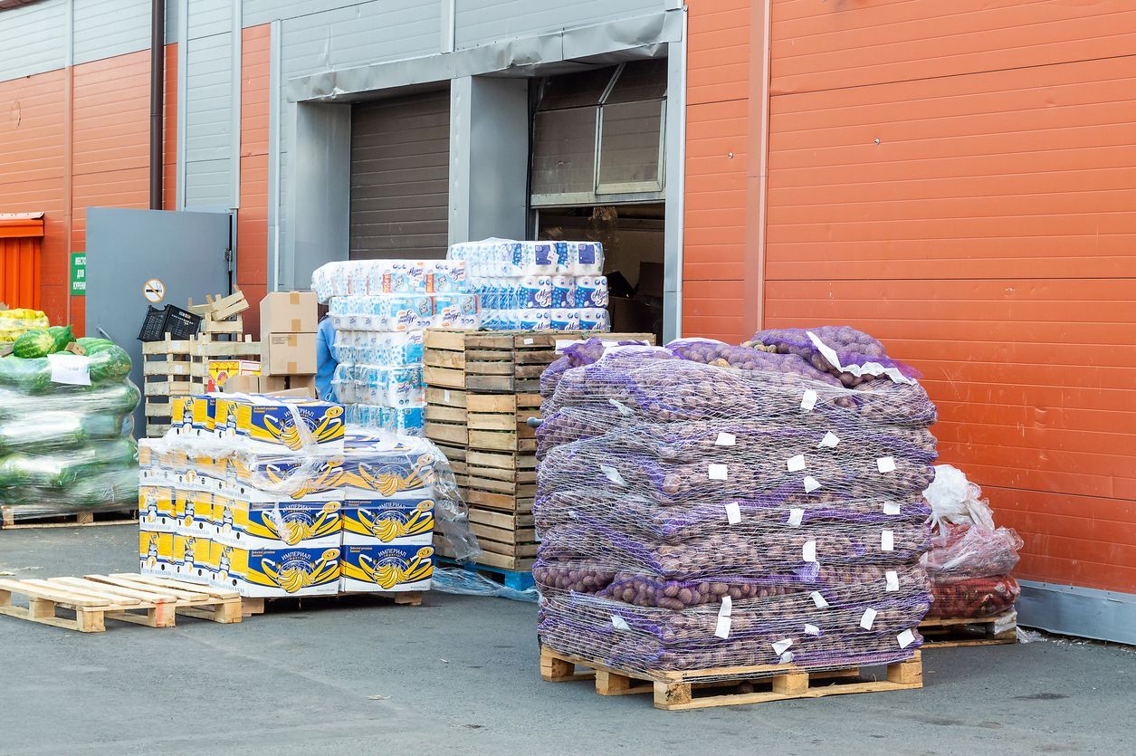 Pallets of produce outside a warehouse: potatoes, water bottles, and boxed goods. A person stands nearby.