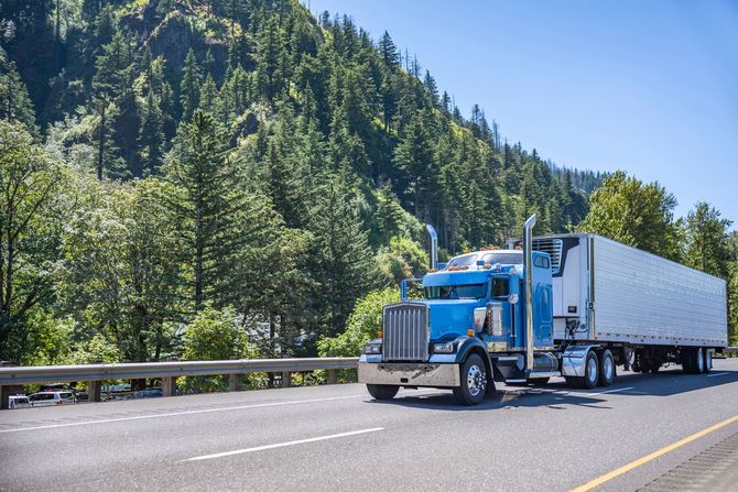 Blue semi-truck driving on a highway with a backdrop of green trees and a blue sky.