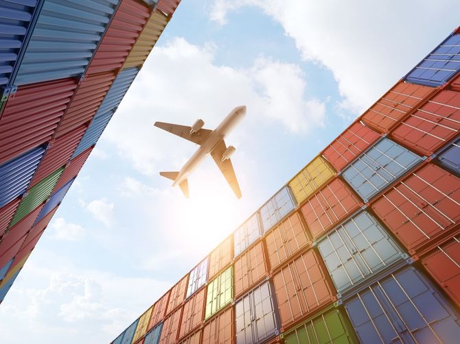 Airplane flying above stacked shipping containers, blue sky.