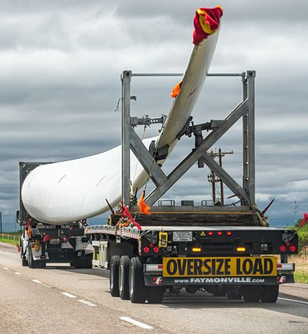 Oversize load truck carrying a large white wind turbine blade on a highway under cloudy skies.