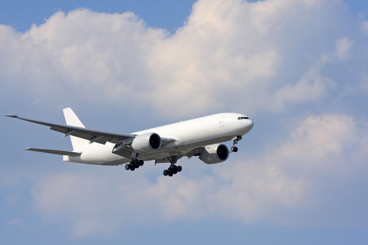 White airplane in mid-air against a blue sky with fluffy white clouds. Landing gear down.