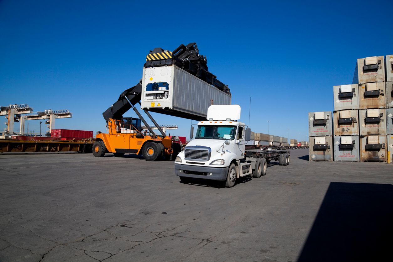 Forklift loading a shipping container onto a white semi-truck in a yard under a clear blue sky.