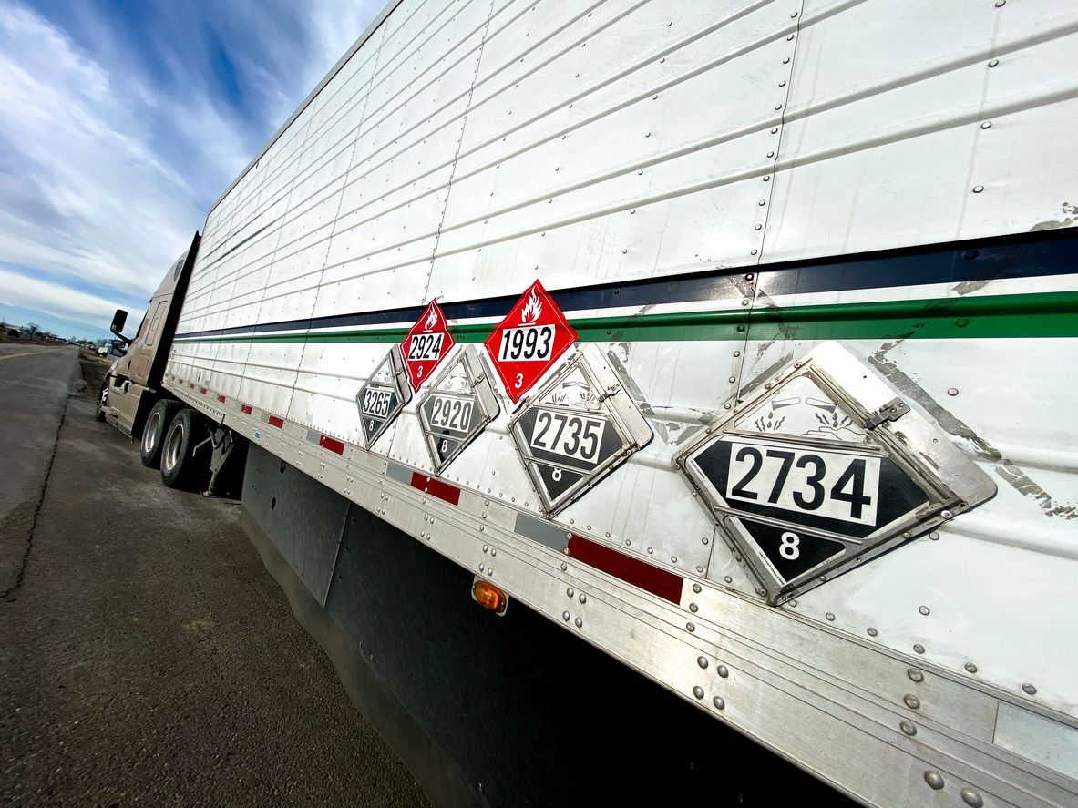 Semi-trailer with hazardous material placards, white and metallic, roadside.