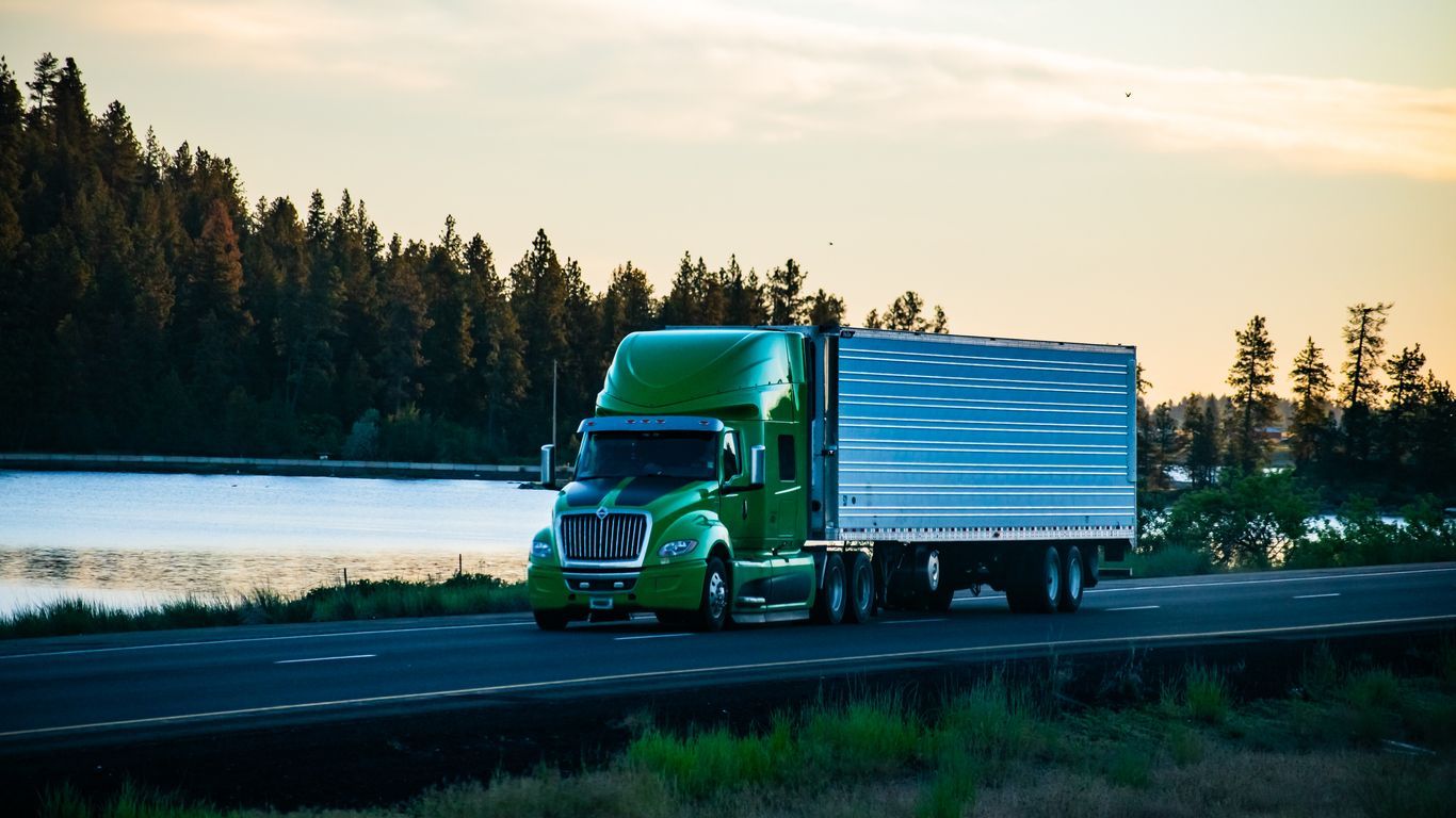 Green semi-truck driving on a road next to a lake with a forest in the background.